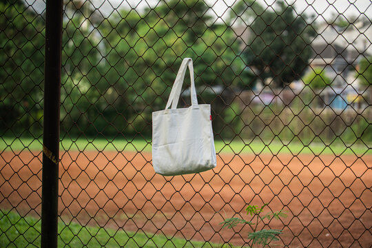 White tote bag hangs on chain-link fence. Soft sunlight highlights fabric's gentle folds. Background features blurred green grass and distant trees. No people visible, suggesting quiet, empty scene