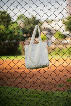 White tote bag hangs on chain-link fence. Soft sunlight highlights fabric's gentle folds. Background features blurred green grass and distant trees. No people visible, suggesting quiet, empty scene