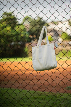 Simple cotton tote hangs on chain-link fence. Sunlight glows behind green trees and grass. Tote shows soft folds and clean lines. Natural scene suggests casual, everyday use