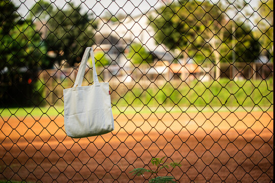Simple cotton tote hangs on chain-link fence. Sunlight glows behind green trees and grass. Tote shows soft folds and clean lines. Natural scene suggests casual, everyday use