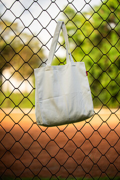 Simple cotton tote hangs on chain-link fence. Sunlight glows behind green trees and grass. Tote shows soft folds and clean lines. Natural scene suggests casual, everyday use