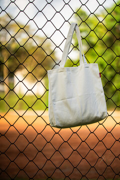 White tote bag hangs on chain-link fence. Blurry background shows green grass and dirt field. Child in distance wears cap and holds bat. Sunlit scene suggests casual park or baseball setting