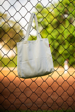 White tote bag hangs on chain-link fence. Blurry background shows green grass and dirt field. Child in distance wears cap and holds bat. Sunlit scene suggests casual park or baseball setting