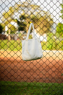 Simple tote bag hangs on chain-link fence. Soft fabric shows subtle wear and natural folds. Background holds green trees and blurred backyard. Scene suggests casual, everyday outdoor setting