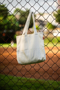 Simple tote bag hangs on chain-link fence. Soft fabric shows subtle wear and natural folds. Background holds green trees and blurred backyard. Scene suggests casual, everyday outdoor setting