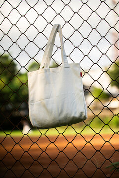 Simple tote bag hangs on chain-link fence. Soft fabric shows subtle wear and natural folds. Background holds green trees and blurred backyard. Scene suggests casual, everyday outdoor setting