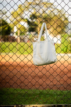 Simple tote bag hangs on chain-link fence. Soft fabric shows subtle wear and natural folds. Background holds green trees and blurred backyard. Scene suggests casual, everyday outdoor setting