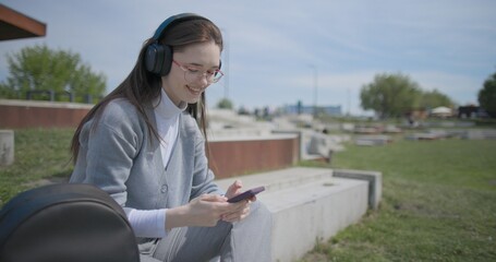 A hand-held shot of a young female student in casual clothes, wearing glasses and headphones, sitting on a concrete in an urban park on a sunny summer day and listening to music from a mobile phone © Anatoly Repin