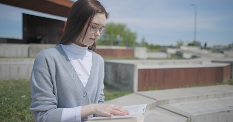 A hand-held shot of a young female student in casual clothes and glasses reading a book while flipping through pages while sitting in an urban park. © Anatoly Repin