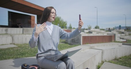 A hand-held shot of a young female student in casual clothes wearing glasses sitting on a concrete bench in an urban park on a sunny summer day, working on a laptop and typing on a keyboard. © Anatoly Repin