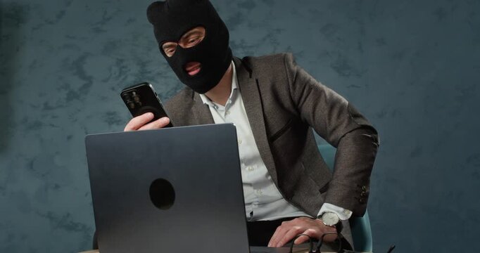 Masked criminal looks at smartphone while working on laptop during scam operation. Fraudster checks phone screen continuing online deception using computer at desk