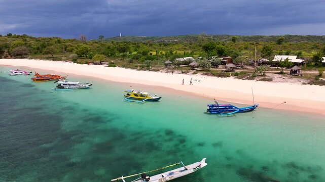 Aerial view of Pink Beach with turquoise water, outrigger boats anchored near the shore, and people walking on the pink sand in Jerowaru, West Nusa Tenggara, Indonesia.