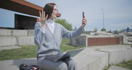 A hand-held shot of a young female student in casual clothes wearing glasses sitting on a concrete bench in an urban park on a sunny summer day, working on a laptop and typing on a keyboard. © Anatoly Repin