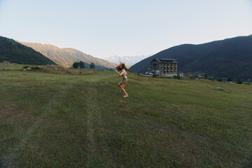 Fototapeta premium Young woman jumps joyfully on green grass field in mountain valley near house during sunny day, enjoying active lifestyle and nature outdoors.