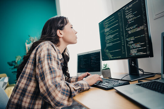Female programmer coding at a home desk with dual monitors, focused software developer writing code on desktop and laptop in a modern workspace