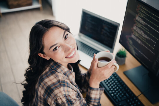 Smiling female programmer taking a coffee break at her home office workstation with laptop and code on screens, relaxed developer working remotely and enjoying a casual productive moment