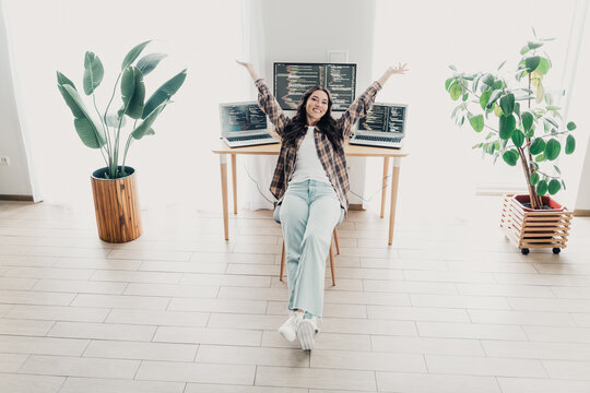 Happy female programmer relaxing in bright home office after coding session with three laptops and plants, celebrating remote work success and comfortable modern workspace