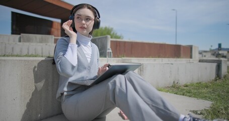 A hand-held shot of a young female student in casual clothes, glasses, and headphones working and typing on a laptop while sitting on a concrete bench in an urban park. © Anatoly Repin