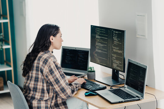 Female software developer working from home, coding on multiple monitors and laptops at a tidy desk while concentrating on programming tasks