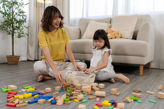 Asian mother teaching daughter good habit through cleaning toys, child placing blocks into basket with guidance, child development, supports discipline and organization skill, early childhood activity