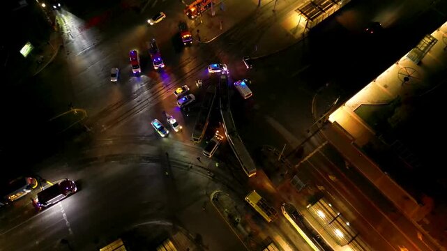 Aerial night view of a busy urban traffic intersection with emergency vehicles, buses, cars, wet reflective pavement, and a brightly lit circular plaza in the background