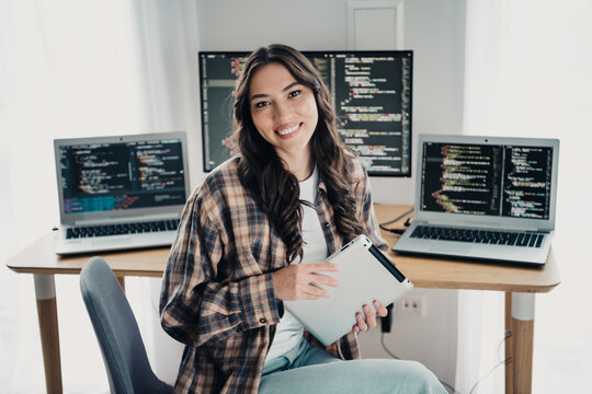 Young female programmer smiling in a bright home office with laptops, monitors showing code and a tablet, working remotely at a casual tech workstation
