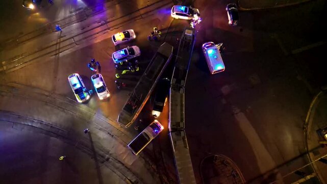 Aerial night view of a busy urban traffic intersection with emergency vehicles, buses, cars, wet reflective pavement, and a brightly lit circular plaza in the background