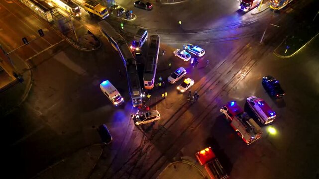 Drone aerial footage of emergency services responding to a tram derailment or traffic accident at a major city intersection during nighttime. Police cars, fire trucks, ambulances, and first responders