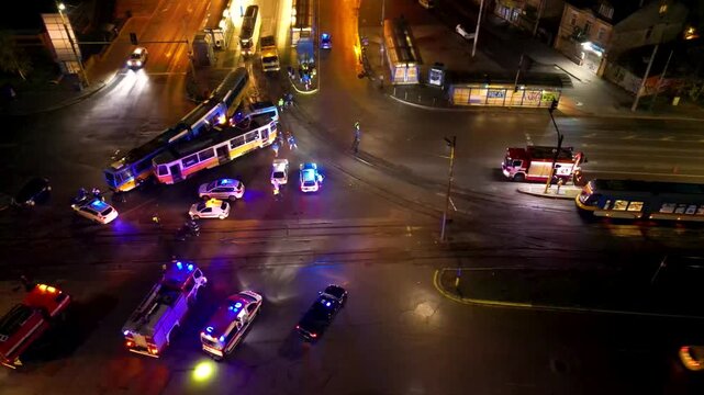 Aerial night view of a busy urban traffic intersection with emergency vehicles, buses, cars, wet reflective pavement, and a brightly lit circular plaza in the background