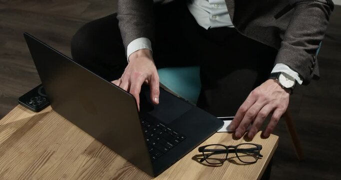 Internet scammer shows bank card while working on laptop at coffee table. Man in suit types messages as presents payment card to support deceptive online scheme