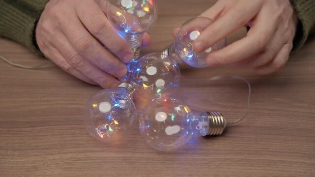 Hands untangling colorful LED string lights shaped like vintage light bulbs on a wooden table.