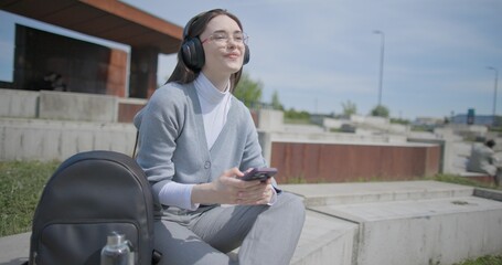 A hand-held shot of a young female student in casual clothes, wearing glasses and headphones, sitting on a concrete in an urban park on a sunny summer day and listening to music from a mobile phone © Anatoly Repin