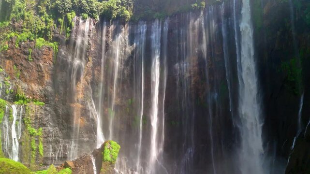 Aerial view of Tumpak Sewu Waterfall with multi-tiered cascades falling down lush green cliffs in Kecamatan Pronojiwo, Jawa Timur, Indonesia.