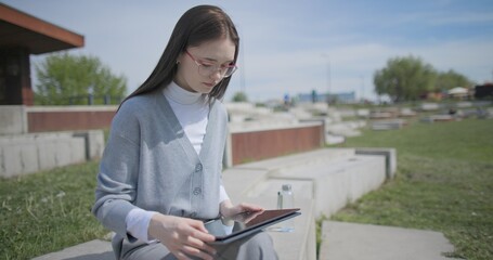 The shot is handheld and shows a young female student in casual clothes wearing glasses sitting on a concrete bench in an urban park and working on a tablet. © Anatoly Repin