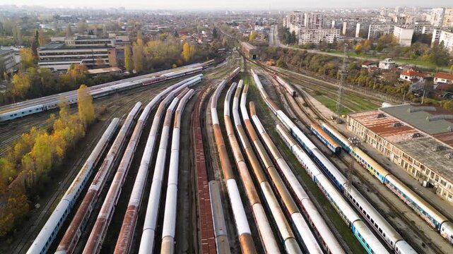 High-angle drone perspective of a sprawling train storage yard with multiple parallel tracks, weathered passenger coaches, maintenance buildings, and nearby residential blocks under soft sunlight