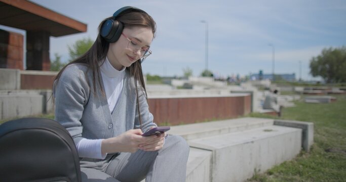 A hand-held shot of a young female student in casual clothes, wearing glasses and headphones, sitting on a concrete in an urban park on a sunny summer day and listening to music from a mobile phone