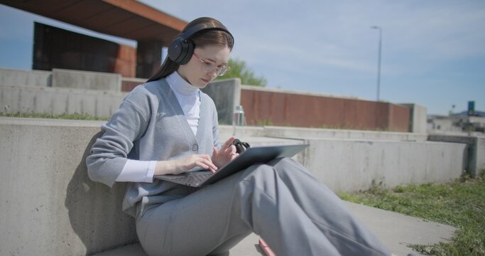 A hand-held shot of a young female student in casual clothes, glasses, and headphones working and typing on a laptop while sitting on a concrete bench in an urban park.