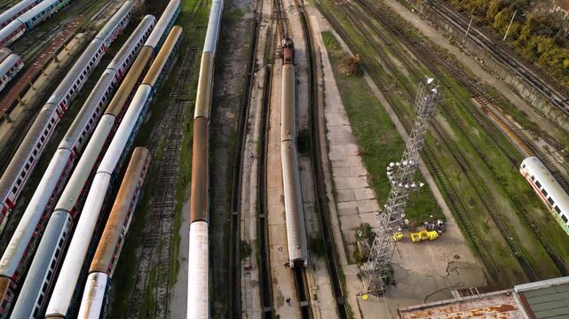 Aerial view of a large railway depot with long rows of parked passenger trains between industrial sheds and city neighborhoods, captured in warm late-afternoon light