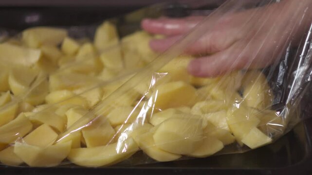 Close-up of female hands placing sliced potatoes into a roasting bag for baking. Home cooking preparation process in a kitchen setting.