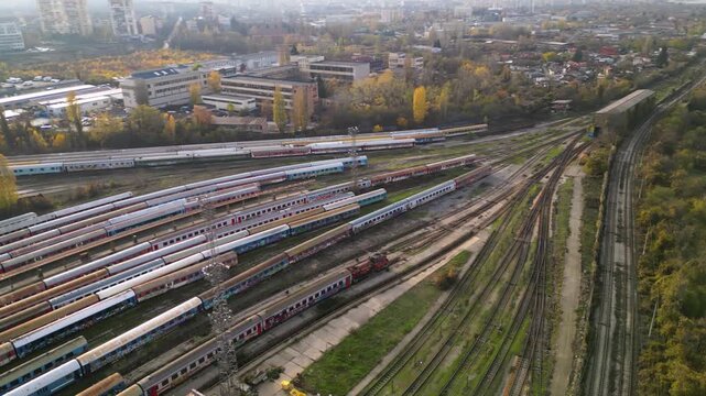 Aerial view of a large railway depot with long rows of parked passenger trains between industrial sheds and city neighborhoods, captured in warm late-afternoon light
