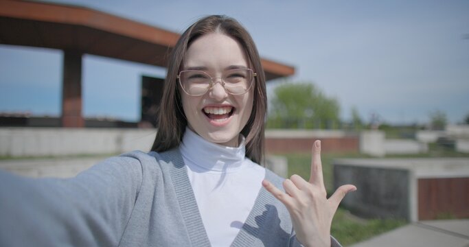 The shot is handheld and shows a young female student in casual clothes wearing glasses, sitting and recording a selfie video on her phone, holding it in her hand and gesturing with her other hand.