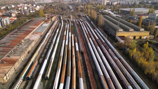 Aerial view of a large railway depot with long rows of parked passenger trains between industrial sheds and city neighborhoods, captured in warm late-afternoon light