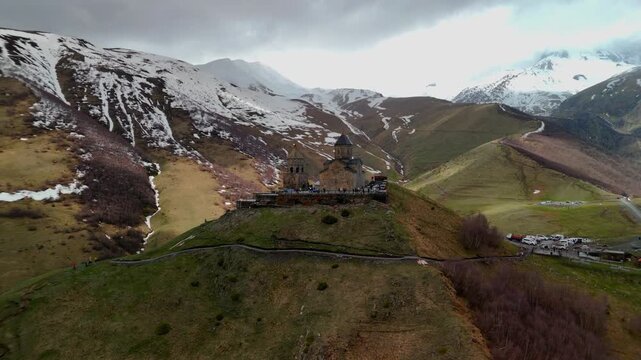 Aerial view of Gergeti Trinity Church standing on a hill against snow-capped mountains and a cloudy sky with people visiting the site Oni, Mtskheta-Mtianeti, Georgia.