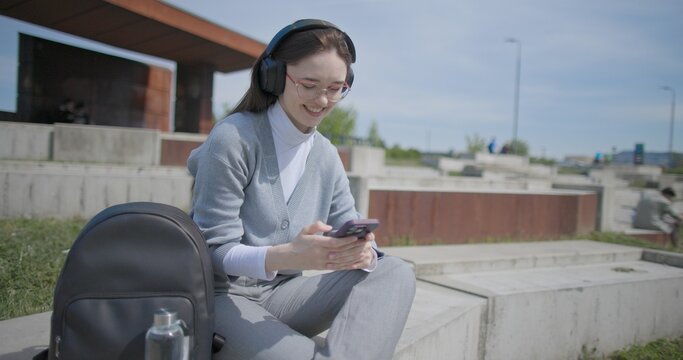 A hand-held shot of a young female student in casual clothes, wearing glasses and headphones, sitting on a concrete in an urban park on a sunny summer day and listening to music from a mobile phone