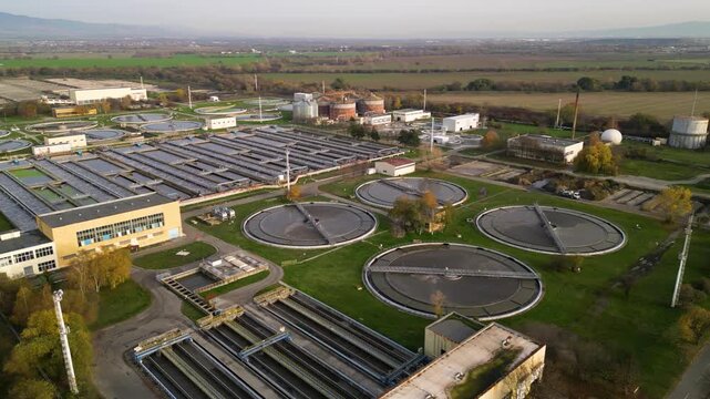 Aerial view of a large wastewater treatment plant with circular settling tanks, rectangular basins, pumping facilities, and green service roads set among agricultural fields and a canal under soft ove