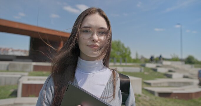 A hand-held portrait of a young female student in casual clothes, wearing glasses and a backpack, holding a notebook and laptop in her hands and posing looking at the camera while standing in a park 