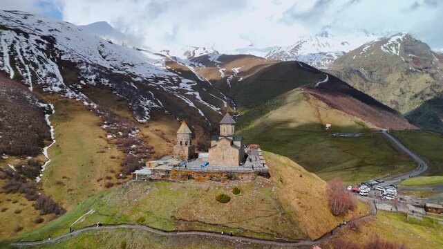 Aerial view of Gergeti Trinity Church on a hilltop surrounded by snow-capped Caucasus Mountains and green valleys in Mtskheta-Mtianeti, Georgia.