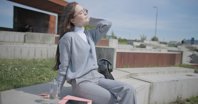 A handheld shot of a young female student in casual clothes sitting on a concrete bench in an urban park, enjoying a sunny summer day.