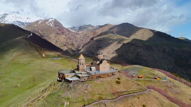 Aerial view of Gergeti Trinity Church on a hilltop with people on the terrace and the Caucasus Mountains in the background, Mtskheta-Mtianeti, Georgia.