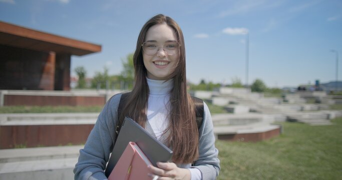 A hand-held portrait of a young female student in casual clothes, wearing glasses and a backpack, holding a notebook and laptop in her hands and posing looking at the camera while standing in a park 